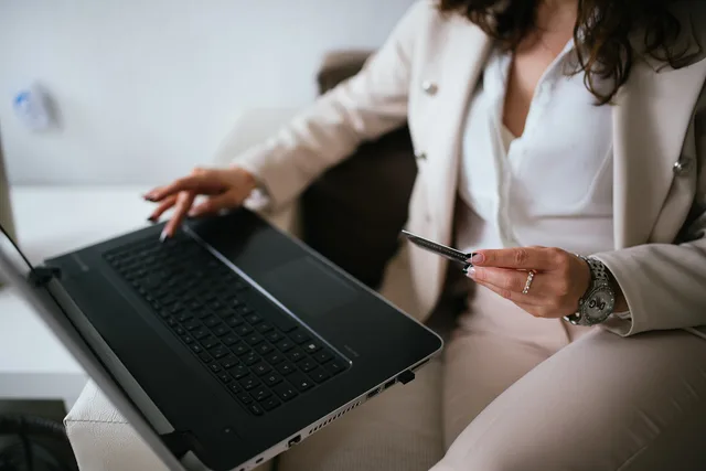 A business professional with curly hair dressed in a suit works on a laptop at home, holding a credit card in one hand and typing with the other, indicating an online shopping activity. Checking shopping links before a seasonal campaign